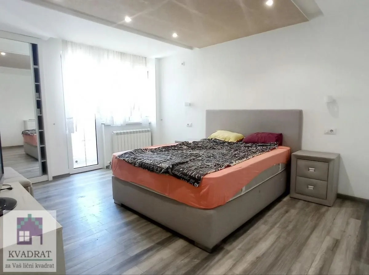 Nameštena spavaća soba sa bračnim krevetom, noćnim stočićem i izlazom na terasu.