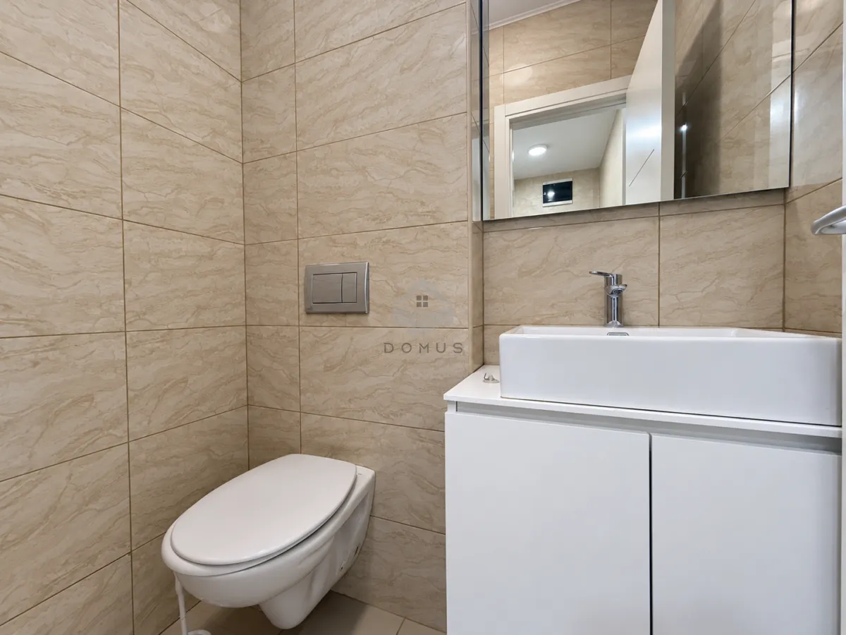 Manje kupatilo obloženo bež pločicama sa ugradnim wc šoljom i bijelim umivaonikom.