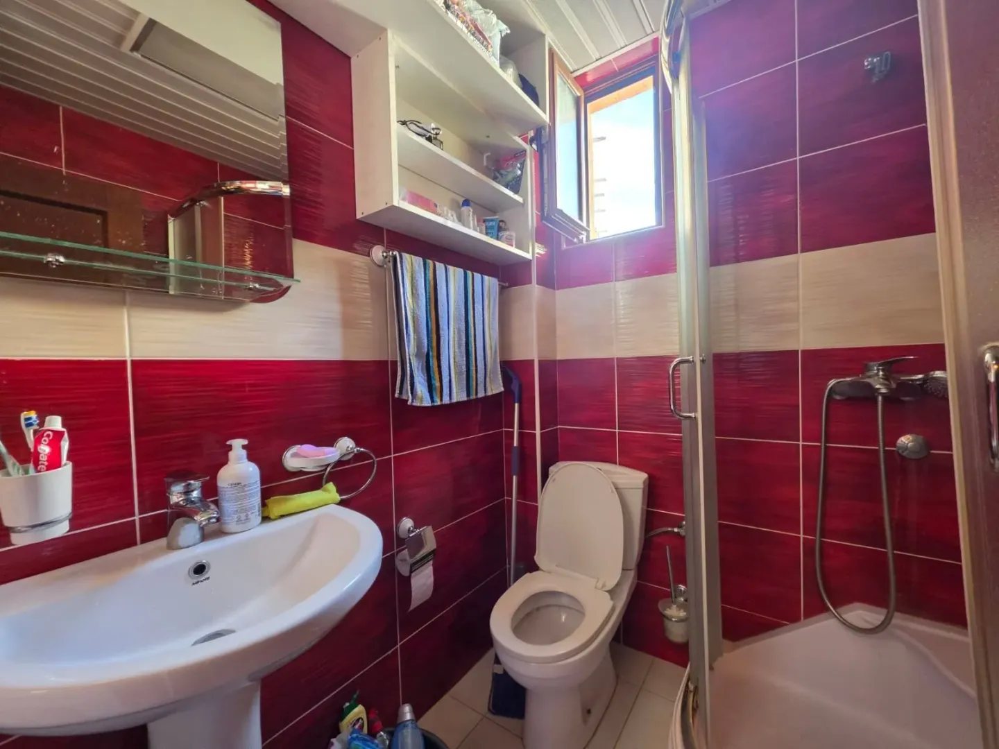 Kupatilo sa umivaonikom, WC šoljom i tuš kabinom u crvenim pločicama.