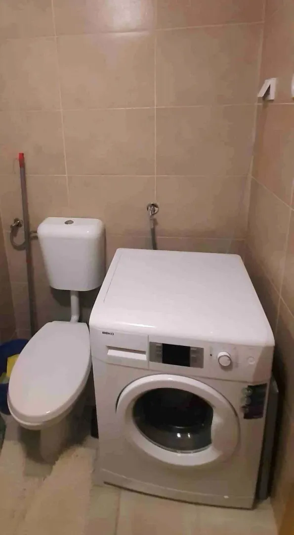 Manje kupatilo sa toaletom i veš mašinom obloženo keramičkim pločicama.