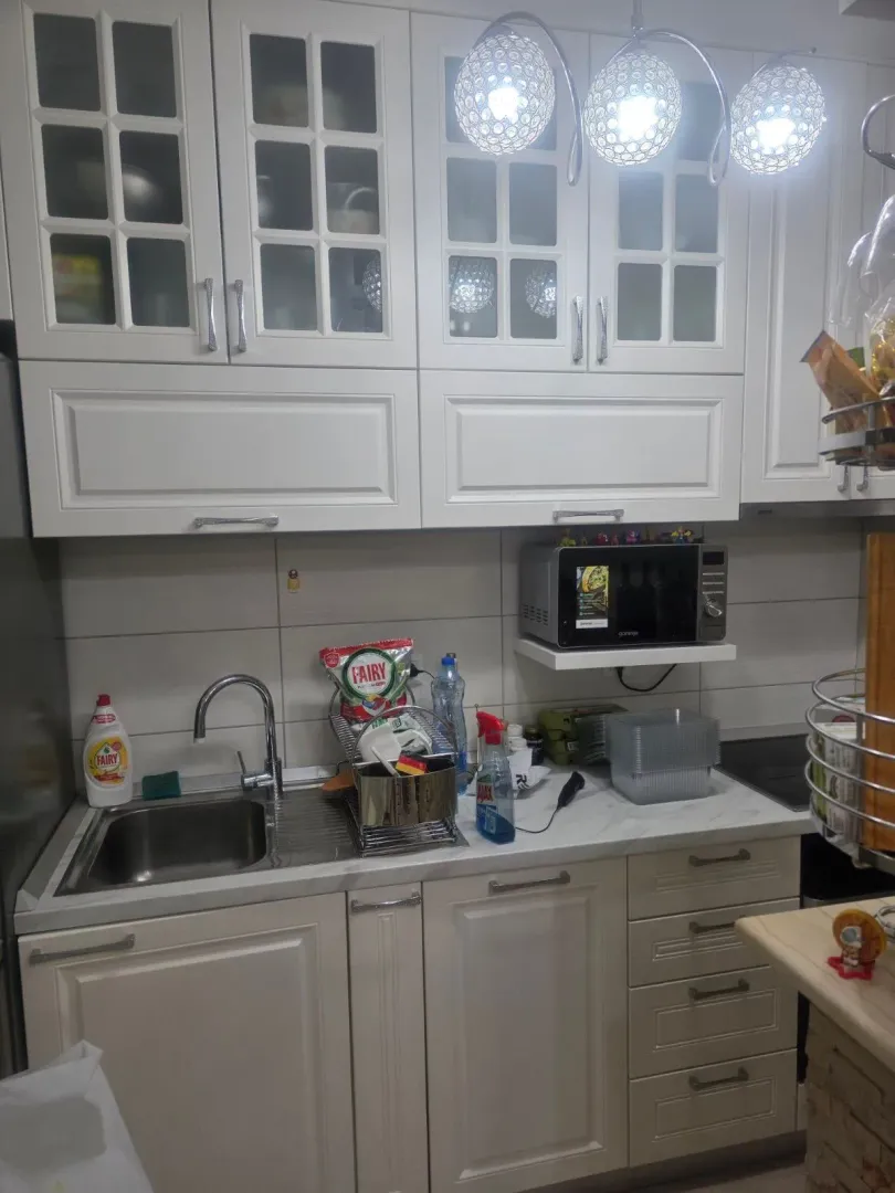 Bijela kuhinja sa sudoperom, radnom površinom i visećim elementima.
