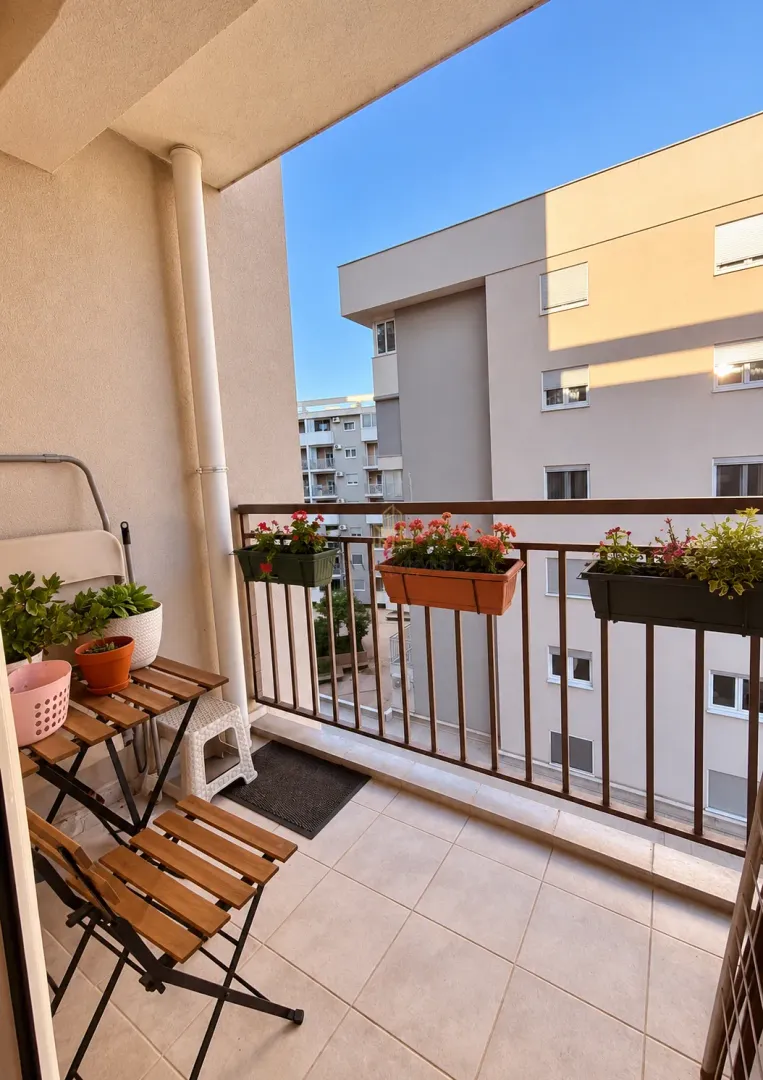 Manji namješteni balkon sa drvenim stolom, stolicama i saksijama sa cvijećem.