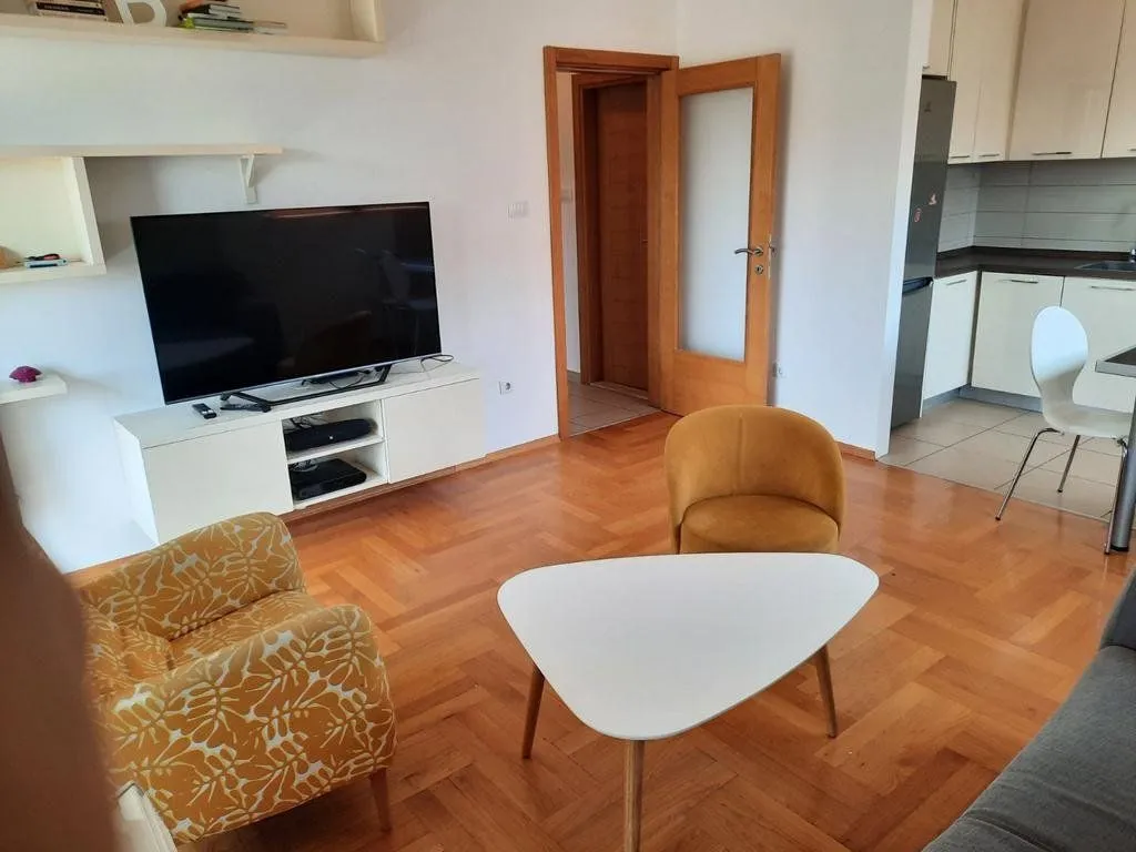 Living room with seating area, television, and a view of the kitchen in the background.