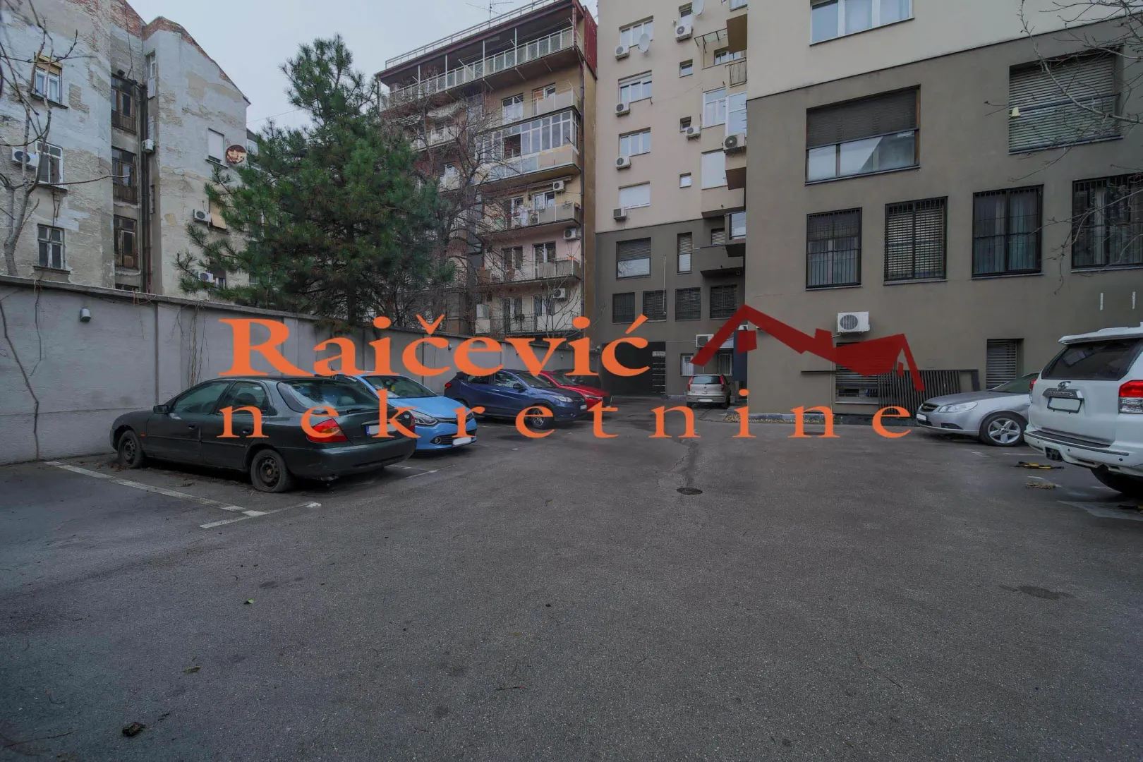 Dvorište sa parking prostorom i zgradama u pozadini.