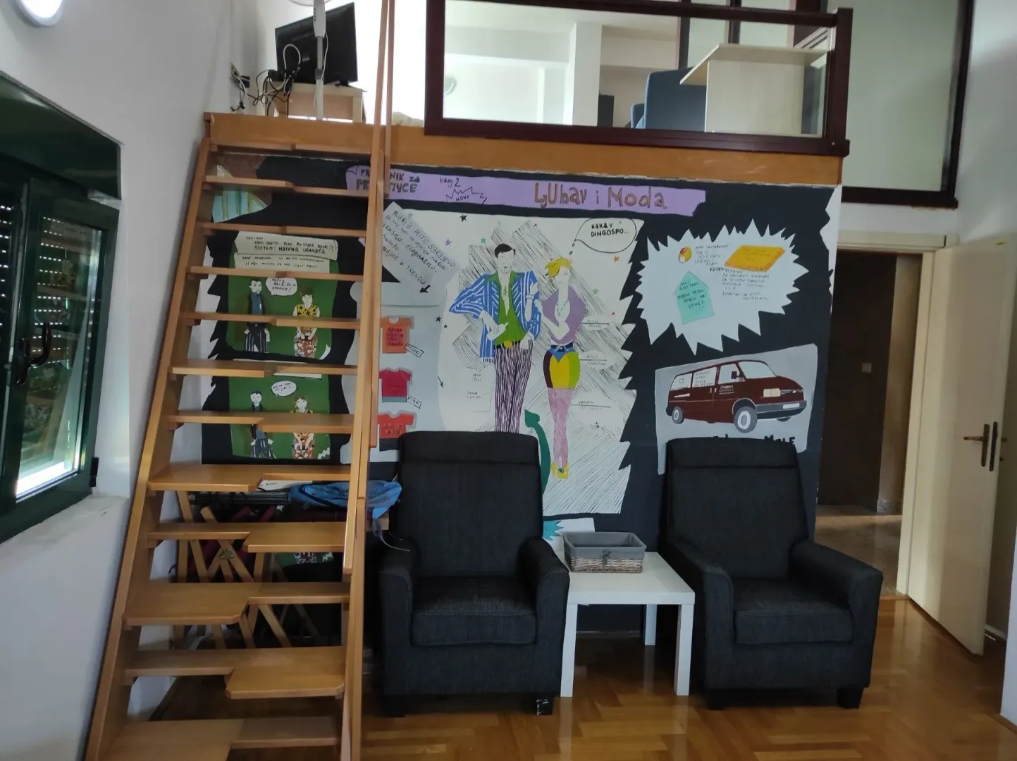 Living room with two armchairs, wooden stairs leading to a loft, and a painted mural on the wall.