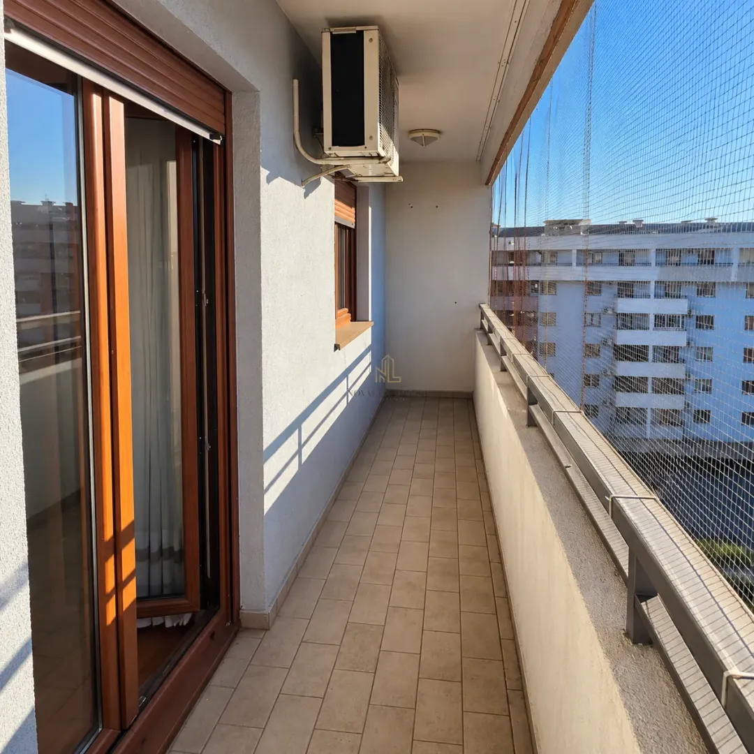 Dugačak popločani balkon sa zaštitnom mrežom i spoljnom jedinicom klima uređaja.