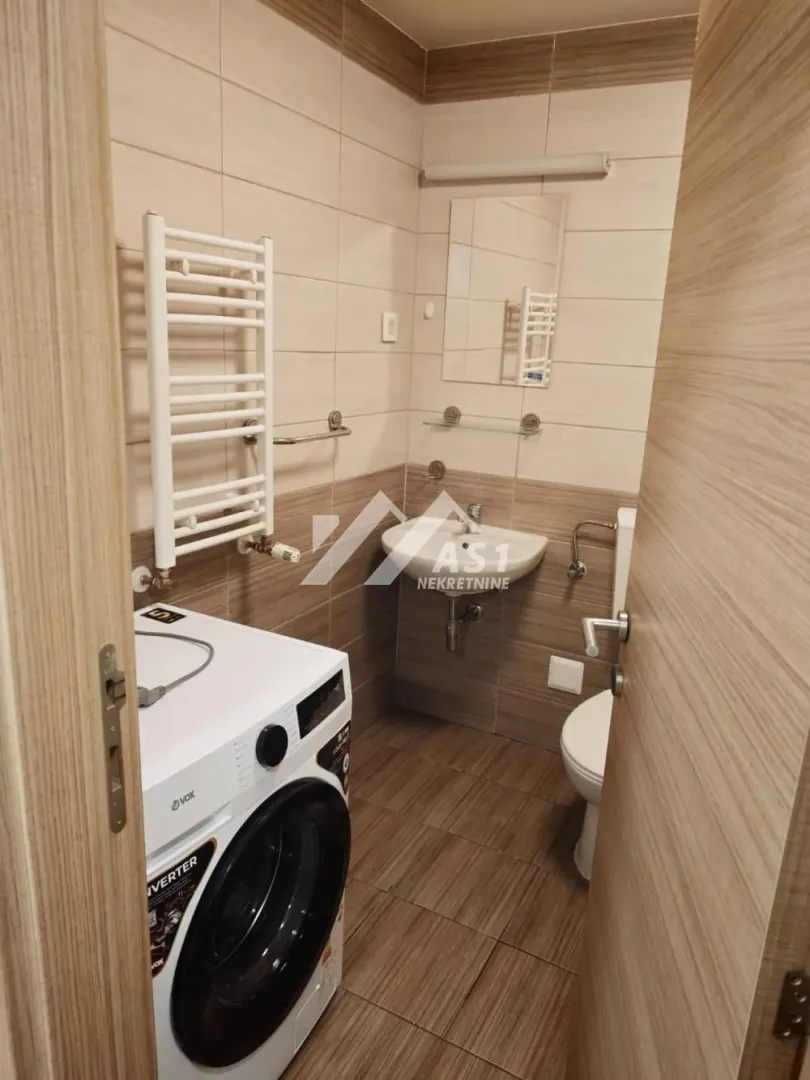 Manje kupatilo obloženo keramičkim pločicama sa veš mašinom, umivaonikom i WC šoljom.