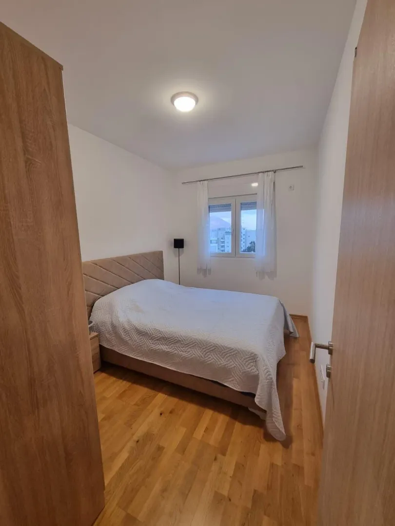 Manja spavaća soba sa bračnim krevetom, drvenim podom i prozorom.