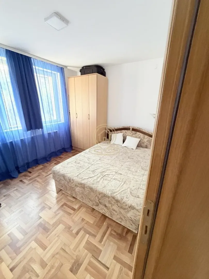 Nameštena spavaća soba sa bračnim krevetom, plakarom i prozorom sa plavim zavesama.
