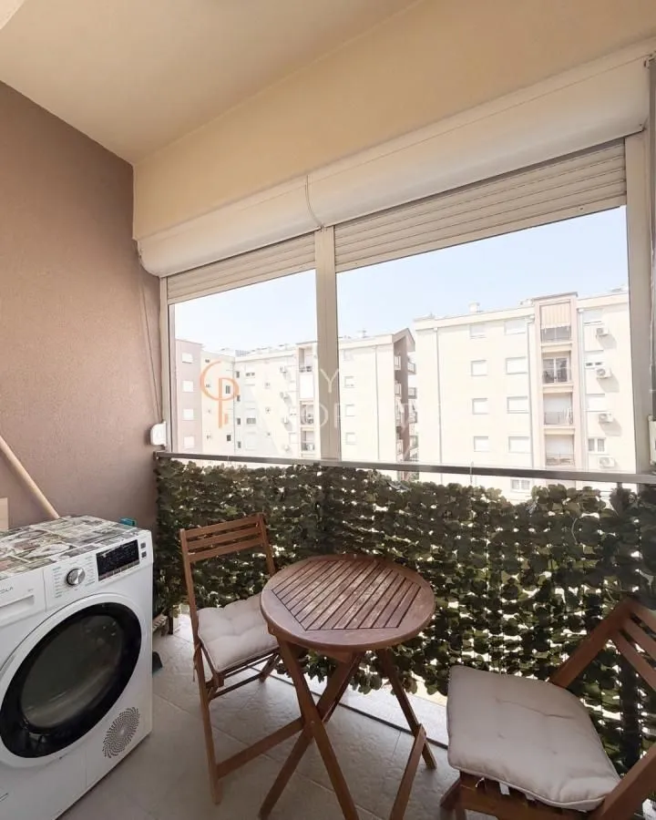 Zastakljeni balkon sa malim stolom, stolicama i veš mašinom.
