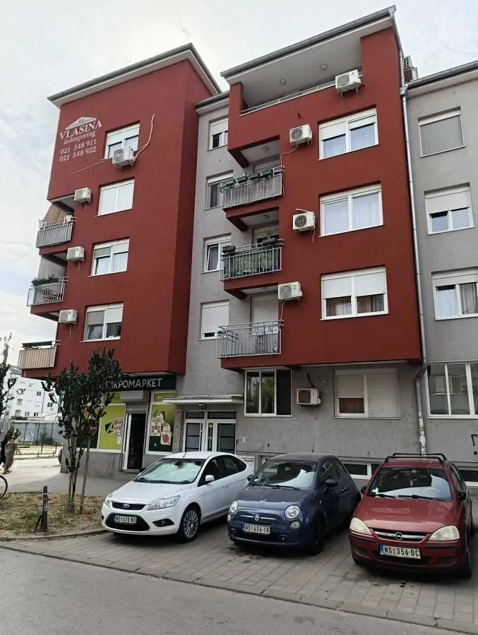Pogled na spoljašnjost stambene zgrade sa parkingom ispred.