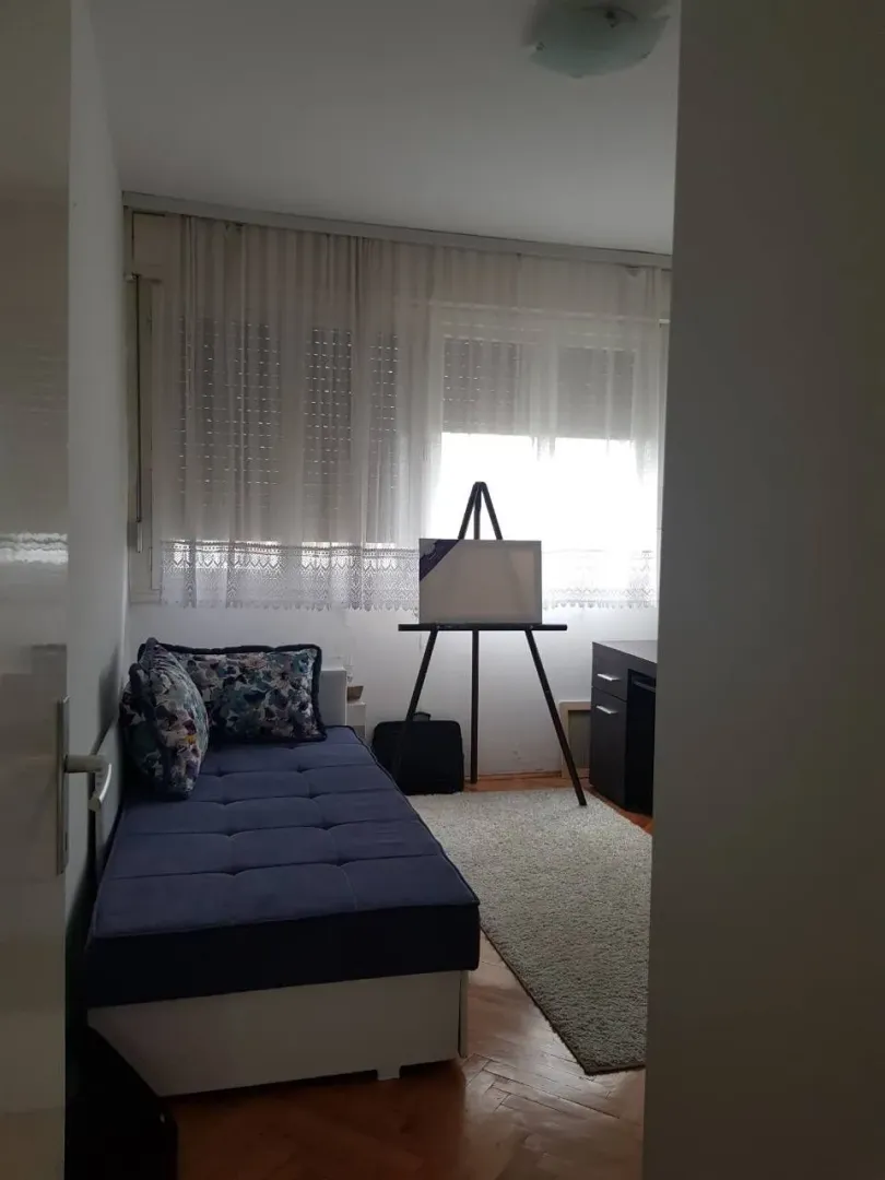 A small bedroom with a blue bed, a desk, and an easel by the window.
