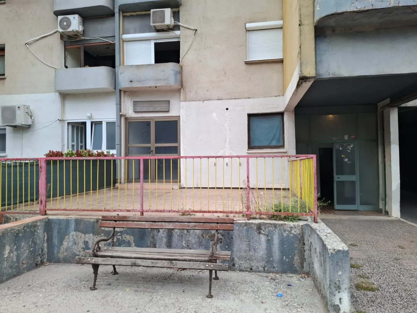 Pogled na ulaz i spoljašnji dio stambene zgrade sa klupom u prvom planu.