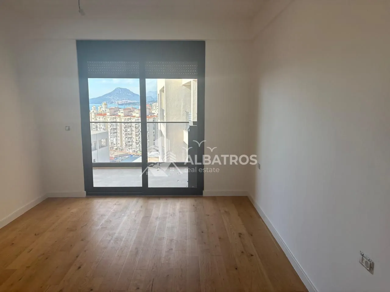 Prazna soba sa drvenim podom i izlazom na balkon sa pogledom na grad.