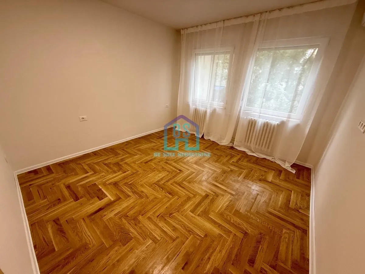 Prazna soba sa parketom i prozorima sa belim zavesama.