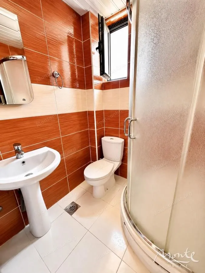 Kupatilo opremljeno tuš kabinom, umivaonikom i toaletom, obloženo narandžastim i bijelim pločicama.