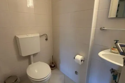 Manje kupatilo obloženo svetlim pločicama sa toaletom i umivaonikom. - image 5