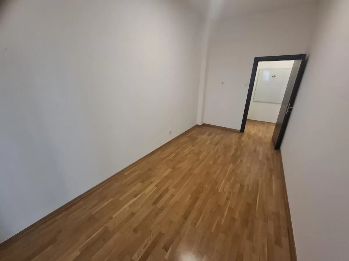 Prazna prostorija sa drvenim parketom i otvorenim vratima koja vode u drugu prostoriju.