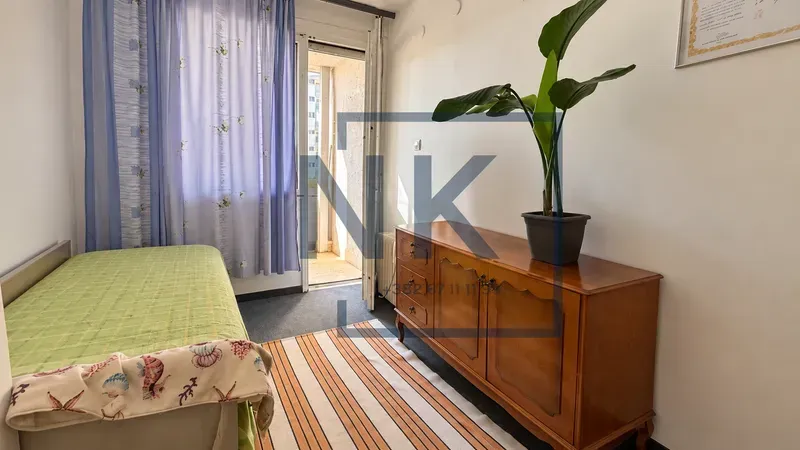 Manja spavaća soba sa krevetom, drvenom komodom i izlazom na balkon.