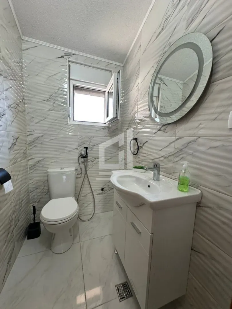 Smaller bathroom with white tiles, washbasin, toilet, and round mirror.