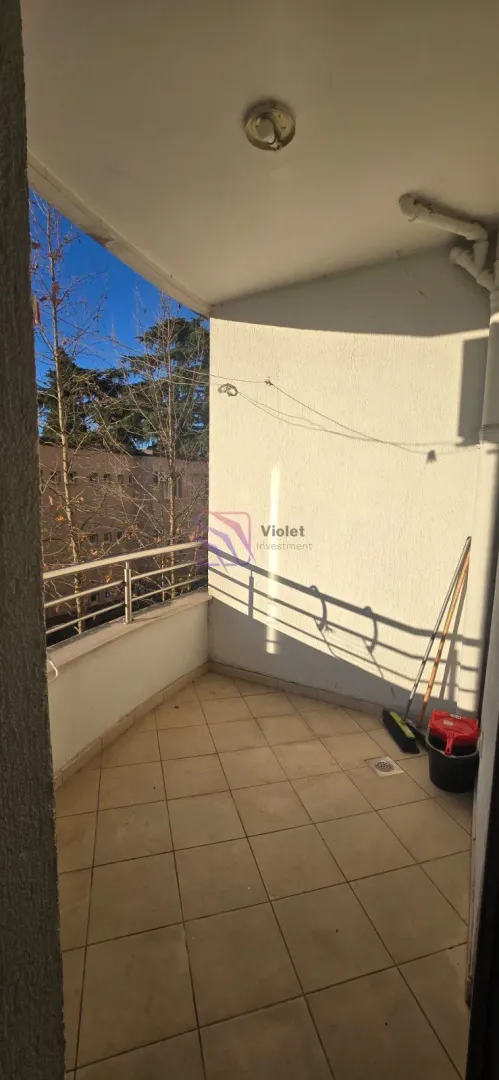 Nenamešten balkon sa pločicama i pogledom na okolne zgrade i drveće.