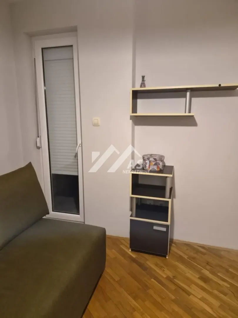 Manja soba sa kaučem, zidnim policama i izlazom na terasu.