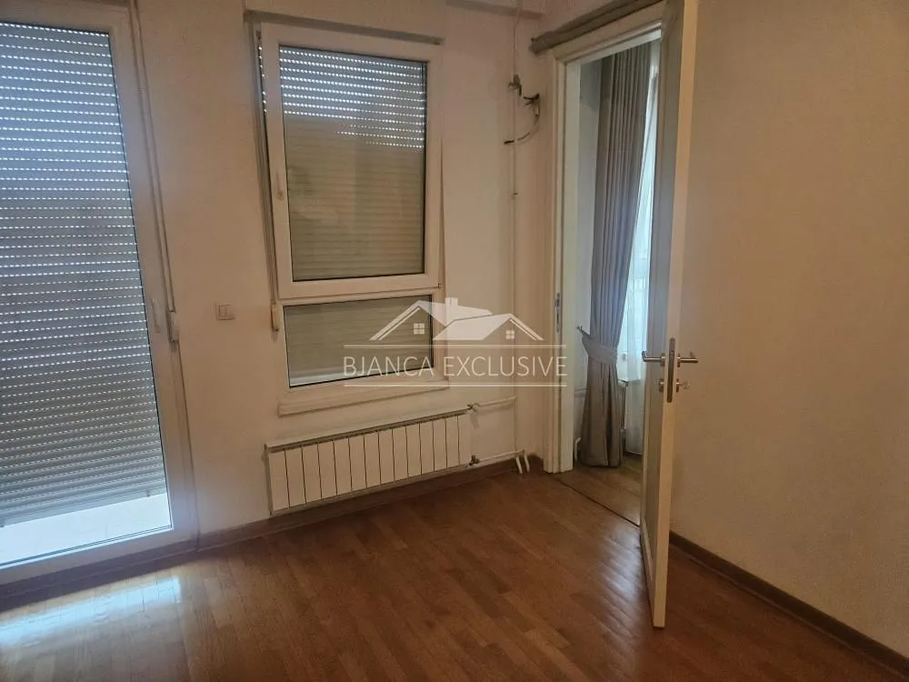 Prazna soba sa drvenim podom, prozorima sa roletnama i vratima koja vode u drugu prostoriju.