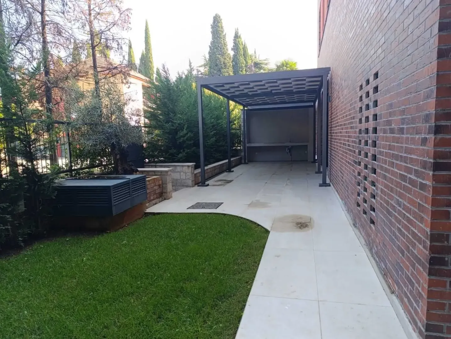Pogled na uređeno dvorište sa popločanom stazom i nadstrešnicom pored kuće od cigle.