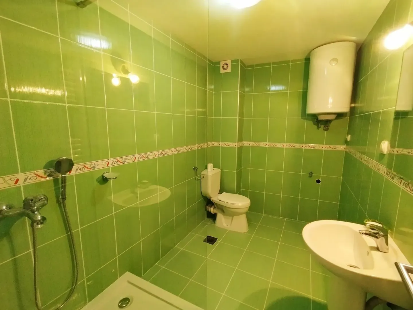 Kupatilo obloženo zelenim pločicama sa tuš kabinom, WC šoljom i umivaonikom.