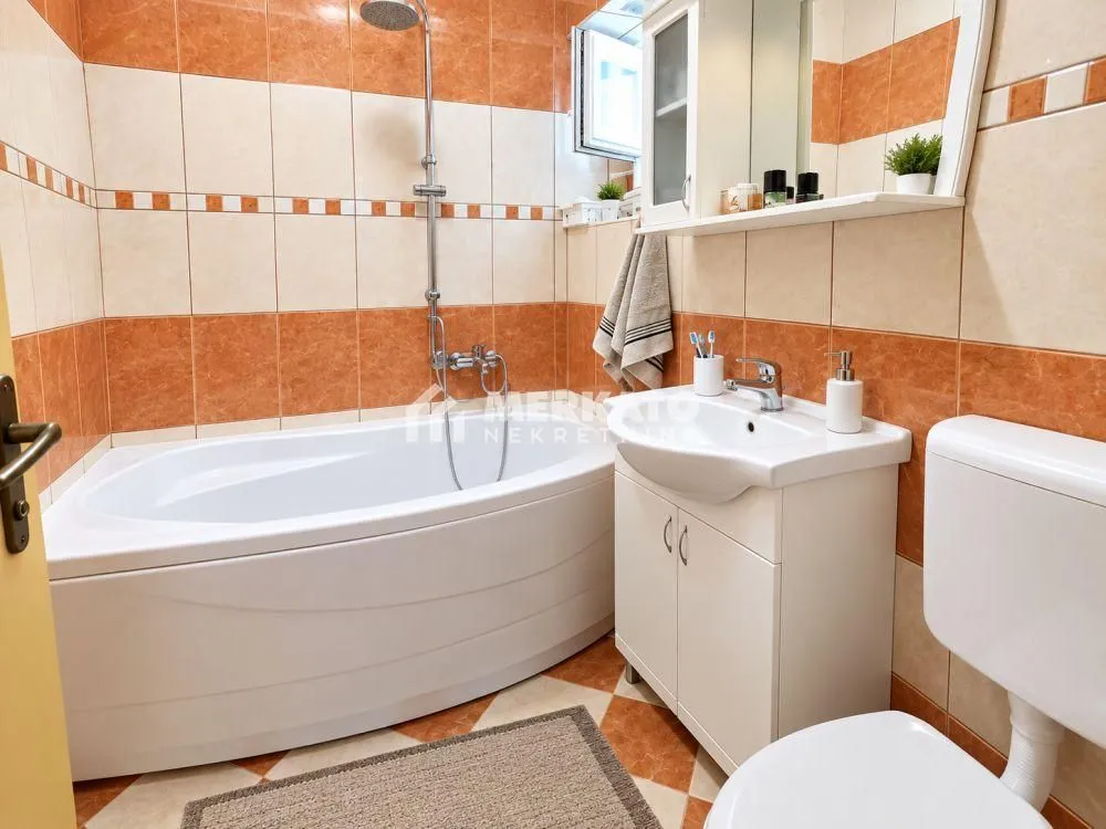 Kupatilo sa kadom, umivaonikom i wc šoljom obloženo keramičkim pločicama u braon tonovima.
