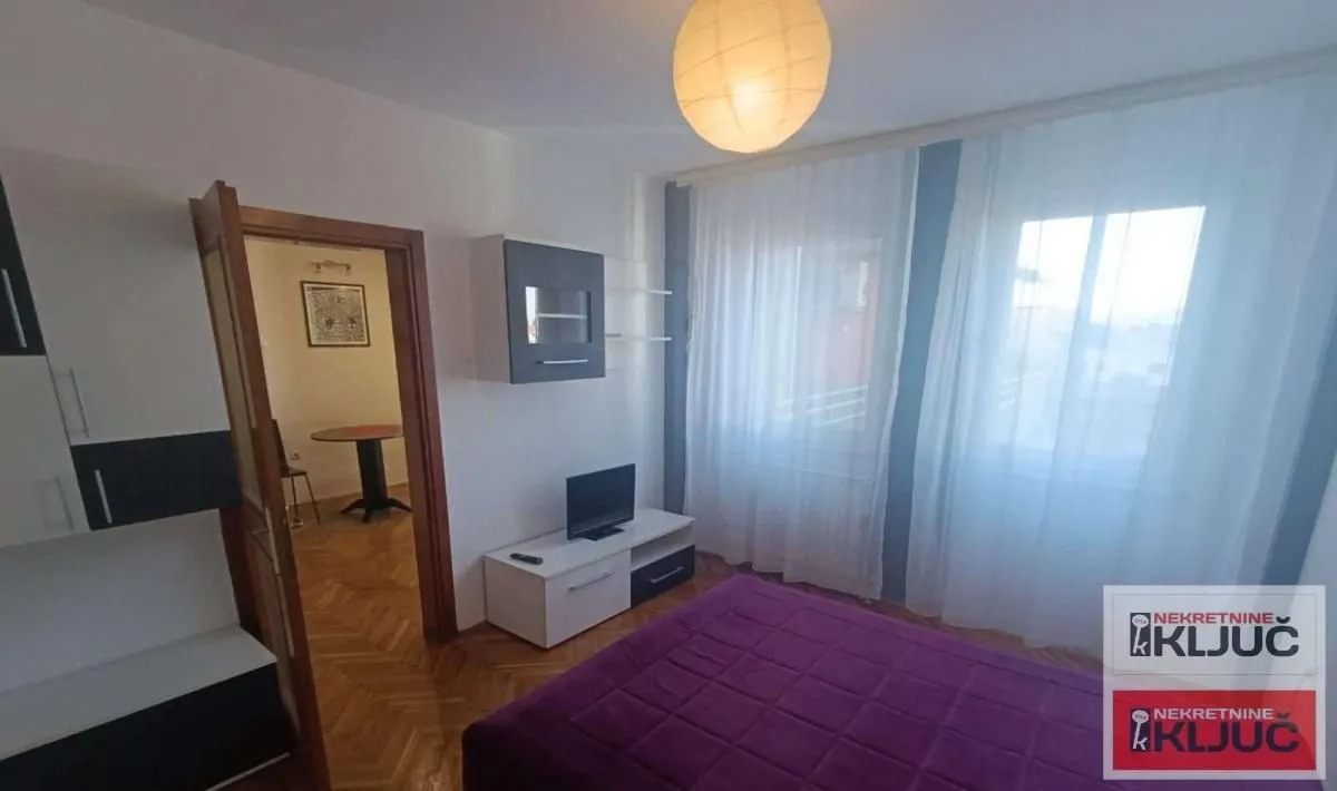 Nameštena spavaća soba sa bračnim krevetom, TV-om i pogledom na prozore sa svetlim zavesama.