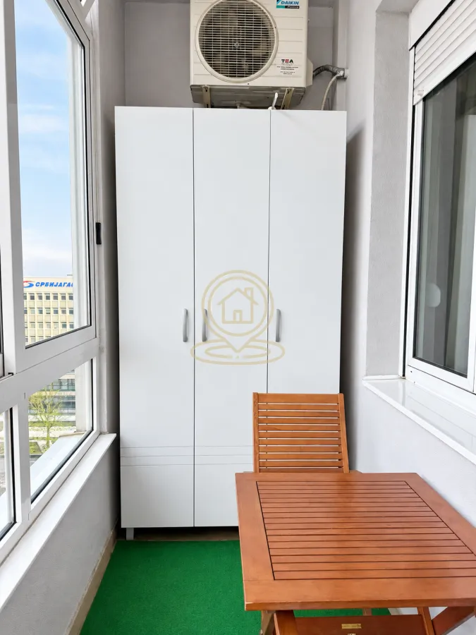 Zastakljeni balkon sa belim garderoberom, drvenim stolom i spoljnom jedinicom klima uređaja.