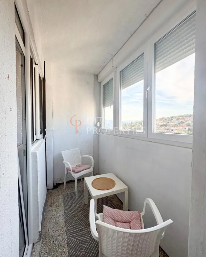Zastakljeni balkon sa malim stolom, dvije stolice i pogledom na grad.