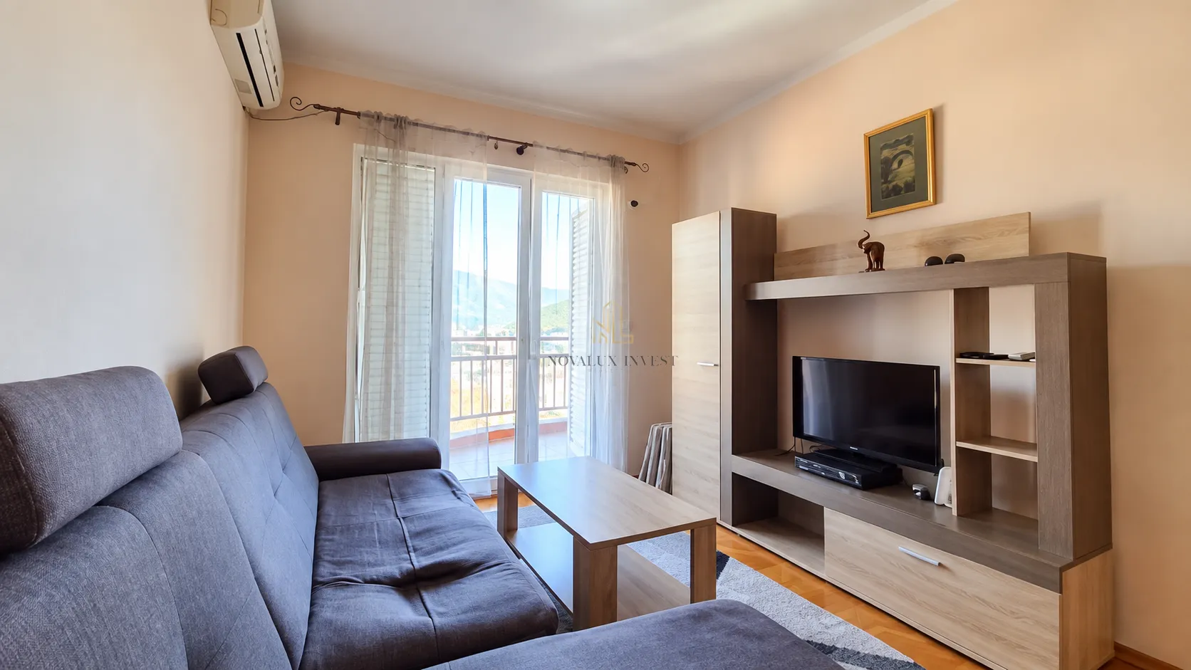 Namještena dnevna soba sa ugaonom garniturom, TV komodom i izlazom na balkon.
