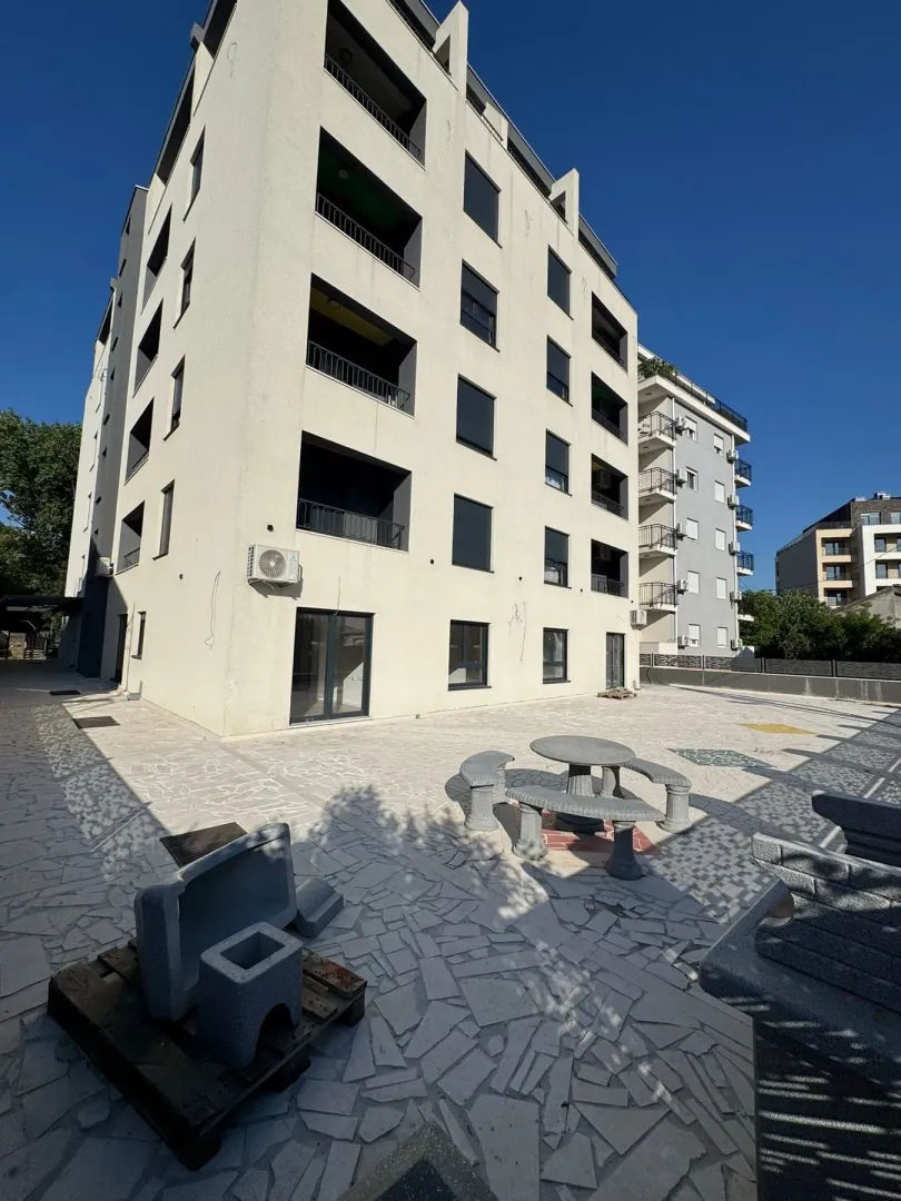 Pogled na spoljašnjost stambene zgrade i popločano dvorište sa klupama.