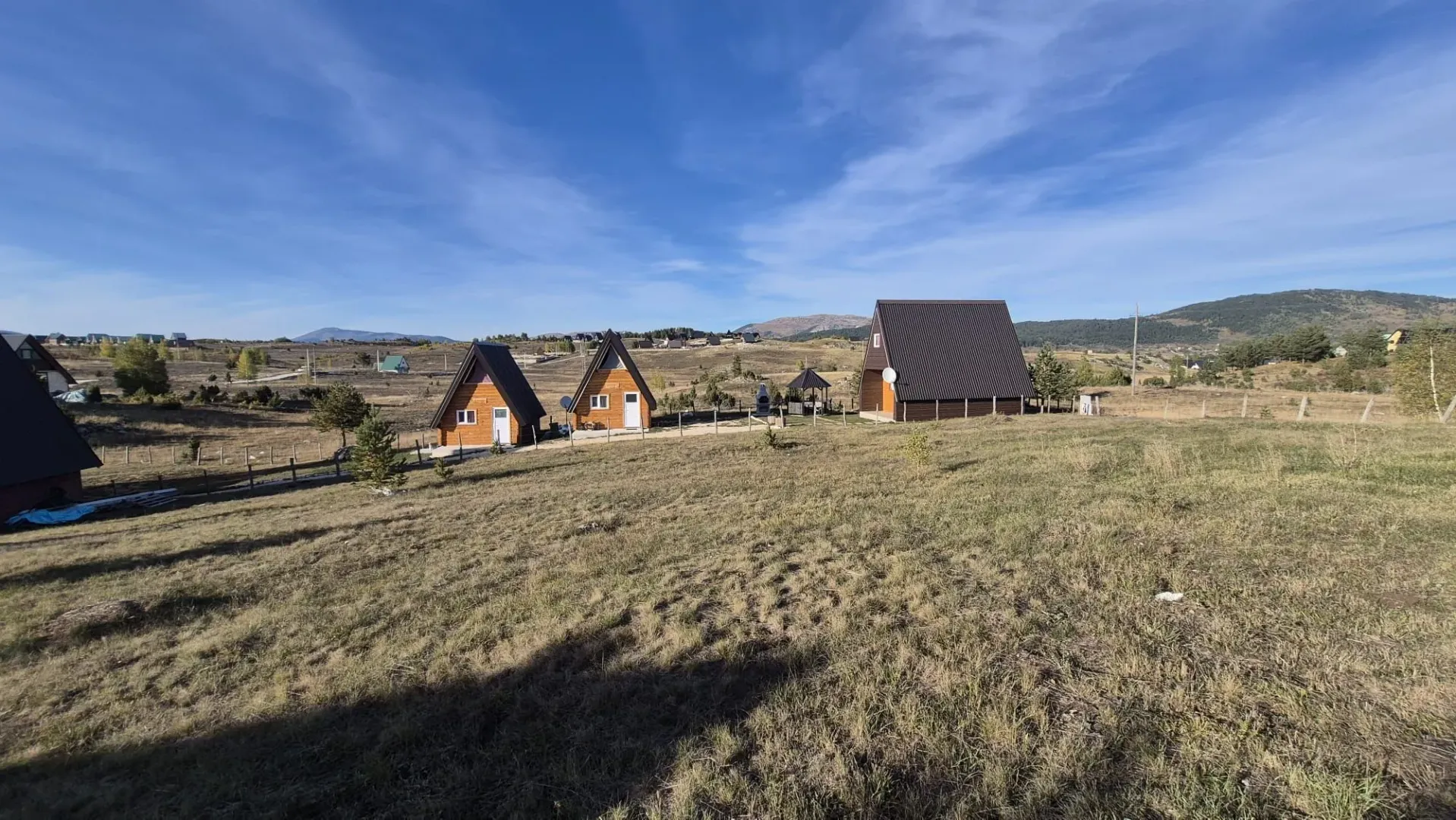 Pogled na drvene vikendice sa prostranim dvorištem i brdima u pozadini.