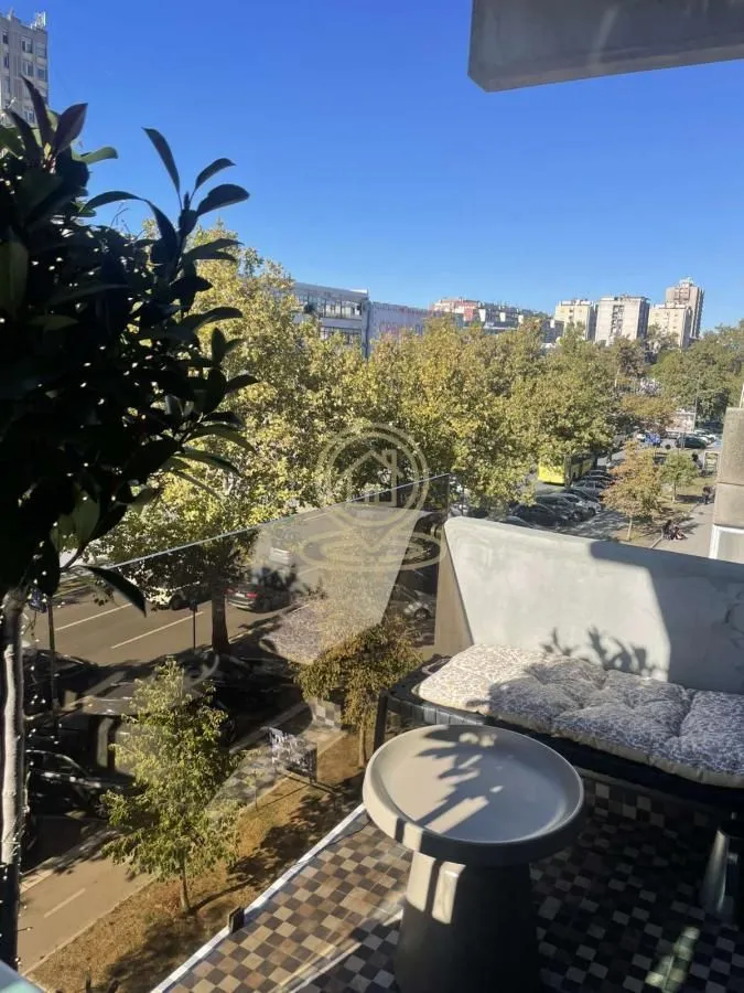 Manji namešten balkon sa garniturom za sedenje i pogledom na gradsku ulicu i drveće.