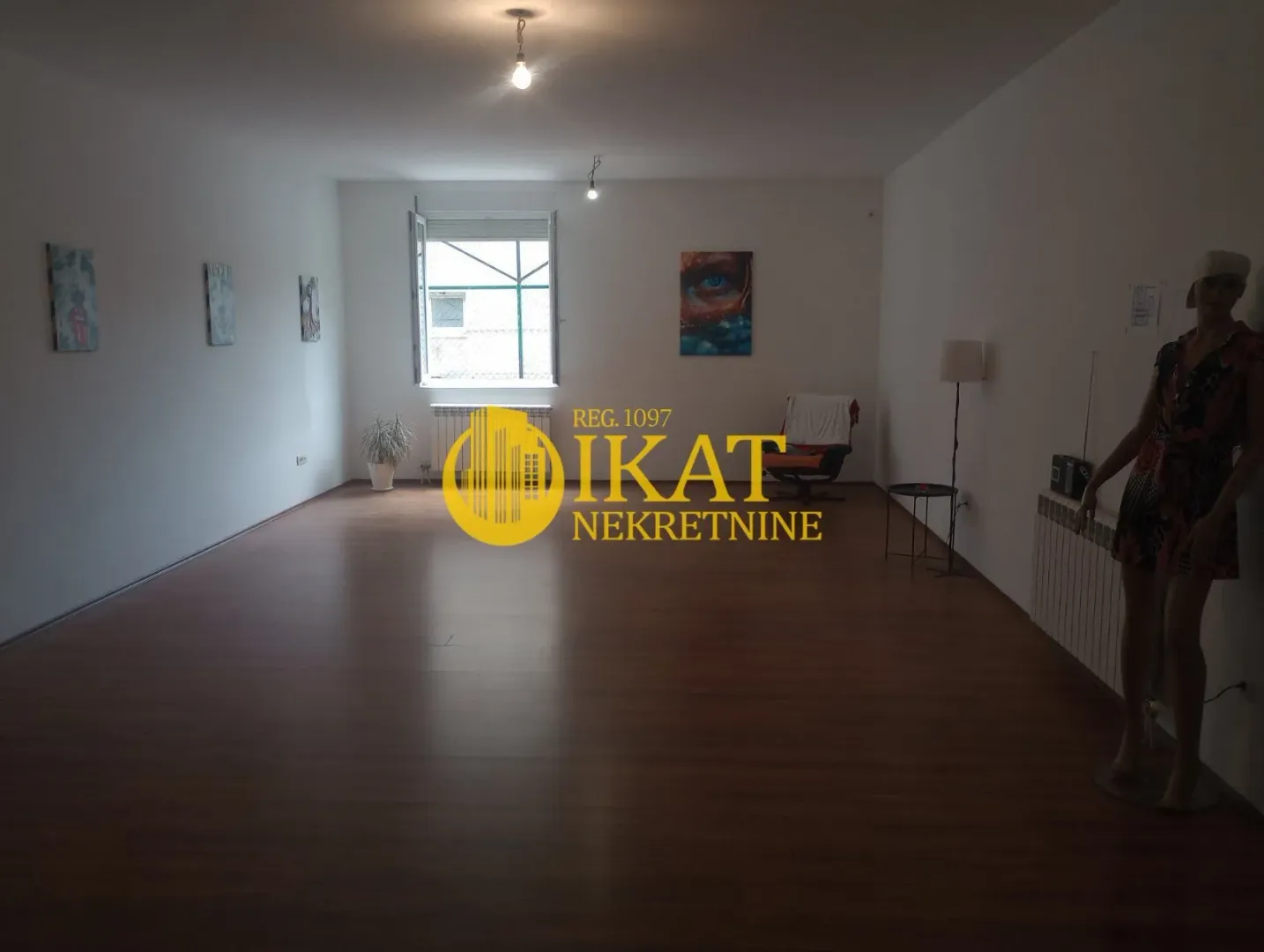 Spacious empty room with wooden flooring, a window, and paintings on the wall.
