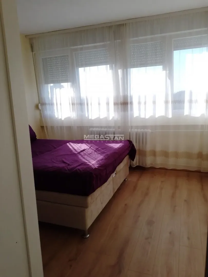 Spavaća soba sa bračnim krevetom i velikim prozorom sa belim zavesama.