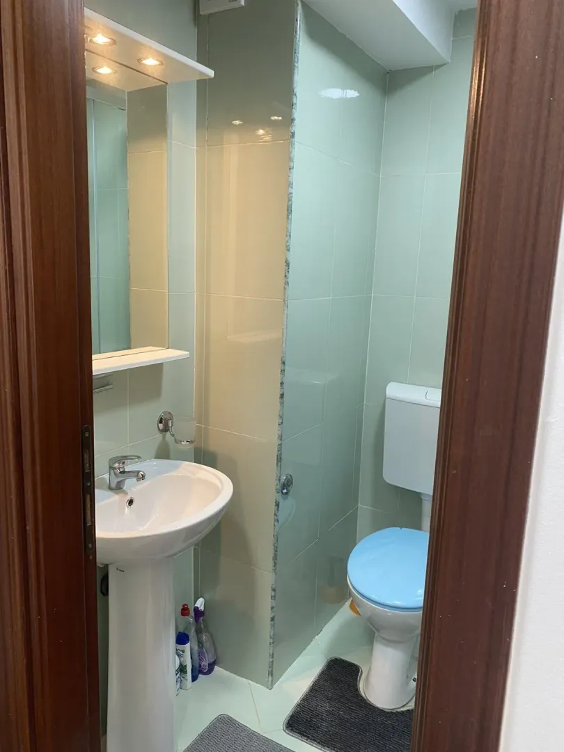 A bathroom with light green tiles, a pedestal sink, and a toilet viewed through a doorway.