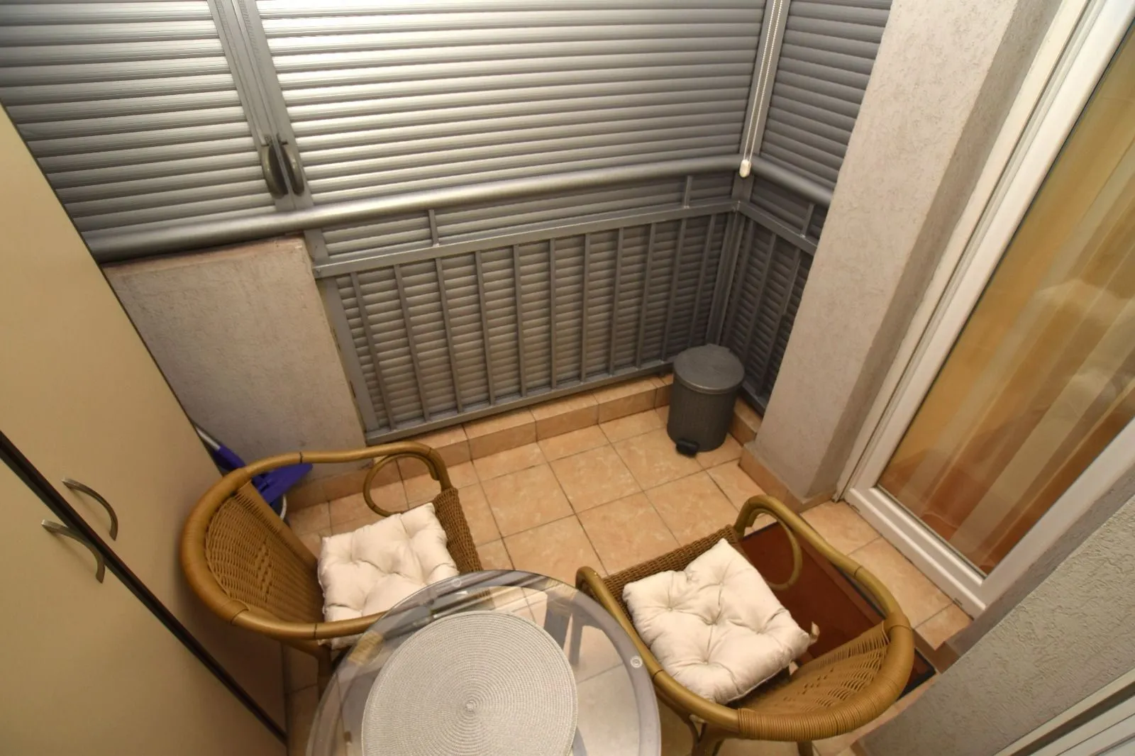 Mali zatvoreni balkon sa dvije pletene stolice i staklenim stolom.