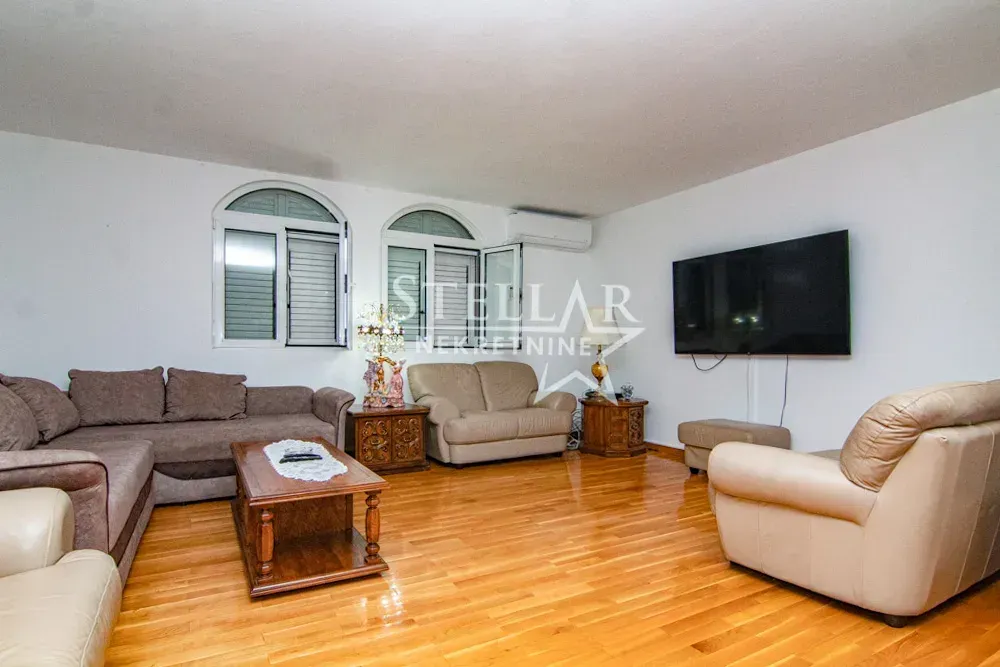 Spacious living room with seating furniture, wooden flooring, and a wall-mounted TV.