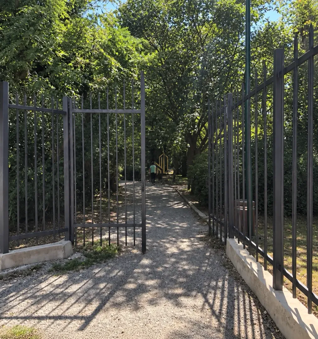 Metalna ograda i otvorena kapija ispred popločane staze i dvorišta sa drvećem.
