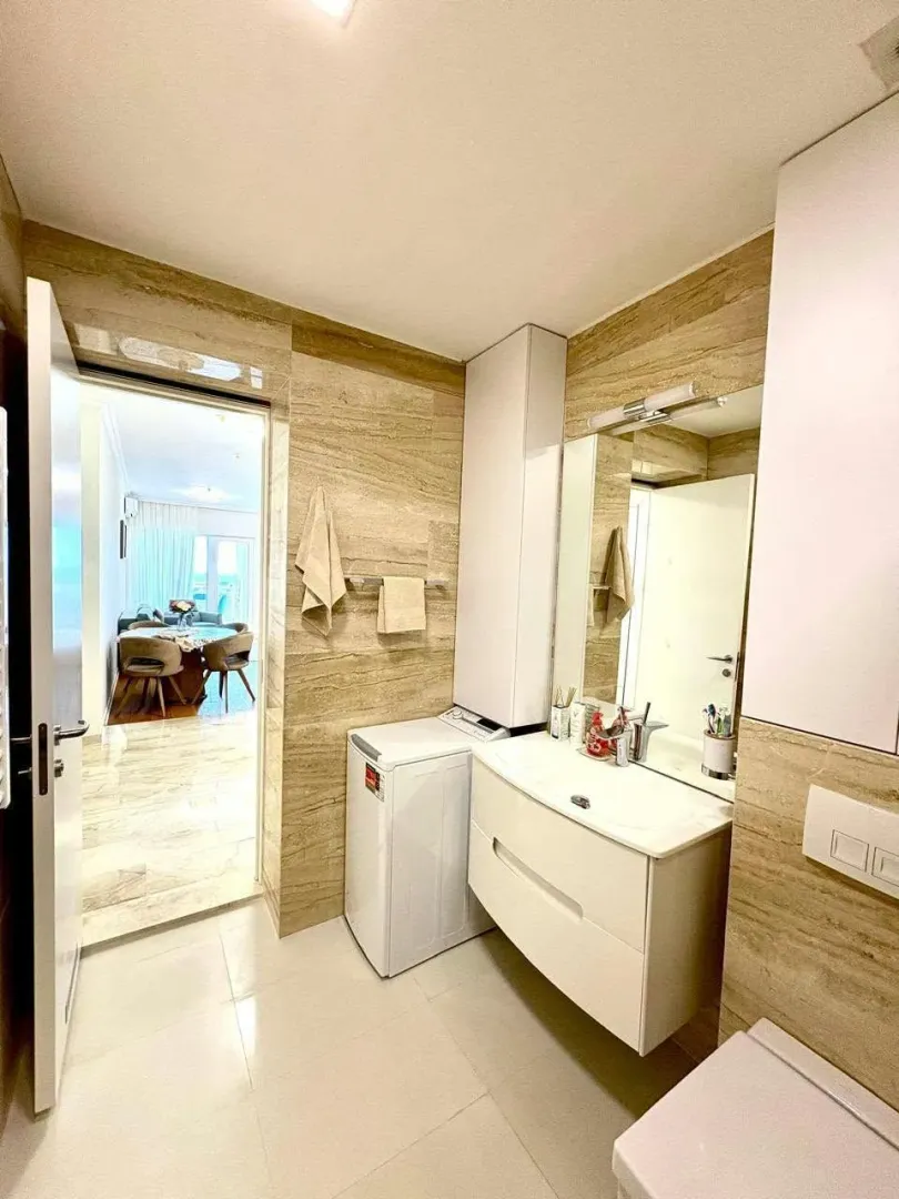 Modernly equipped bathroom with a vanity, washing machine, and a view into the living room.