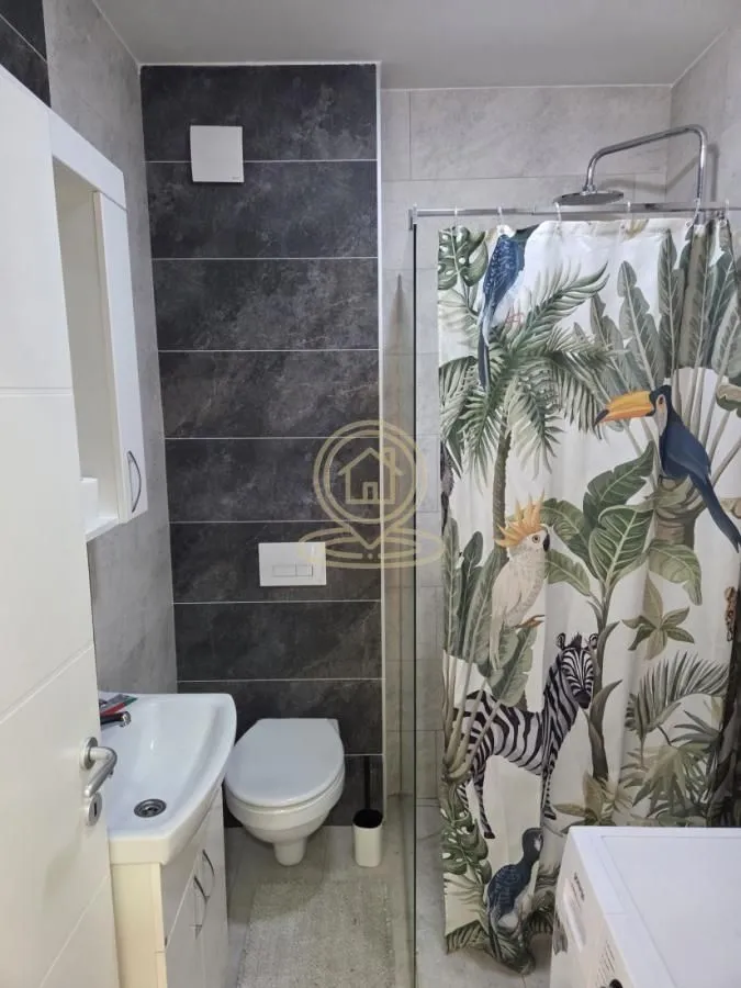 Kupatilo opremljeno umivaonikom, WC šoljom i tuš kabinom sa zavesom.