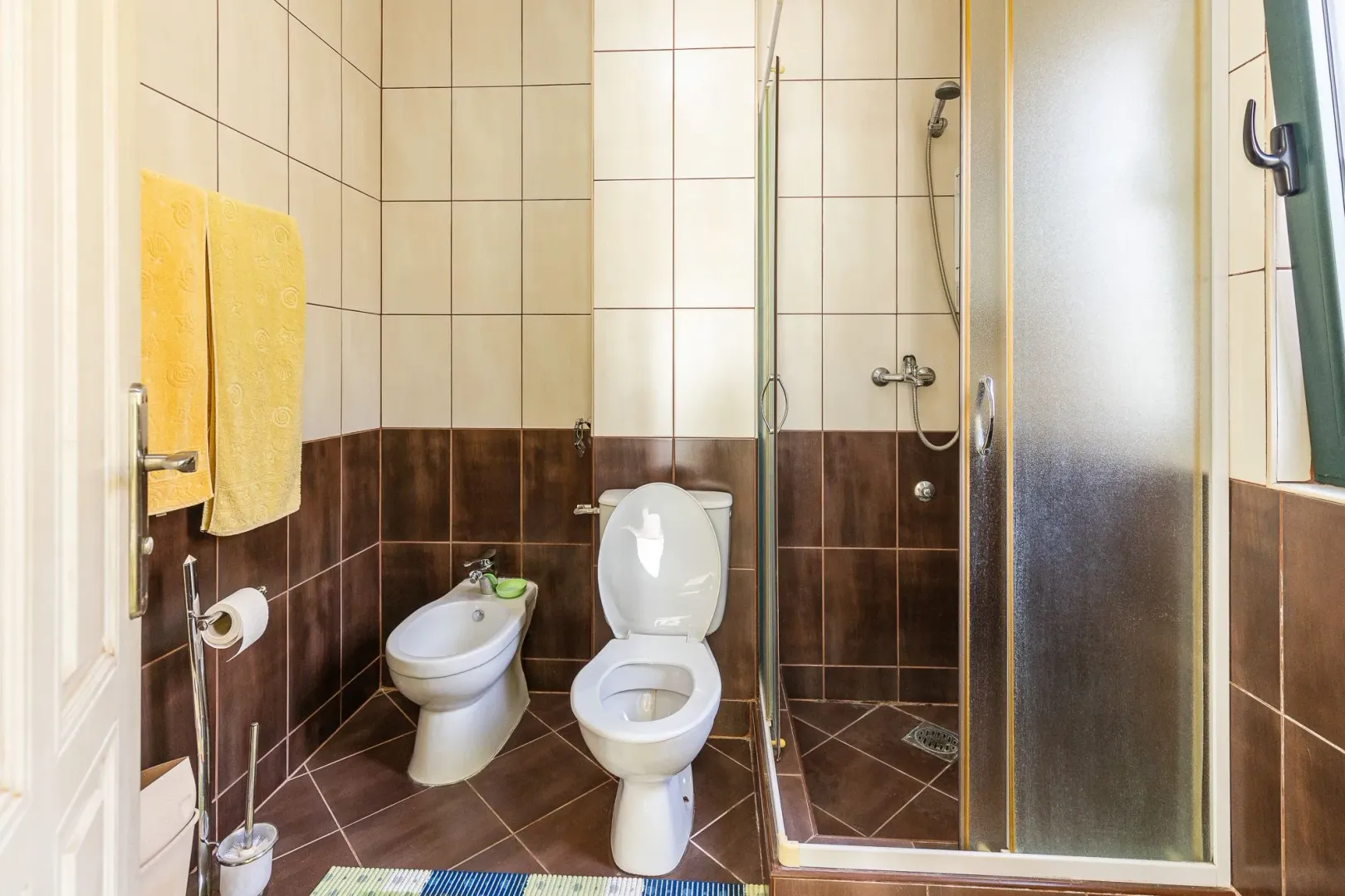 Manje kupatilo sa tuš-kabinom, šoljom i bideom u smeđim i bež pločicama.