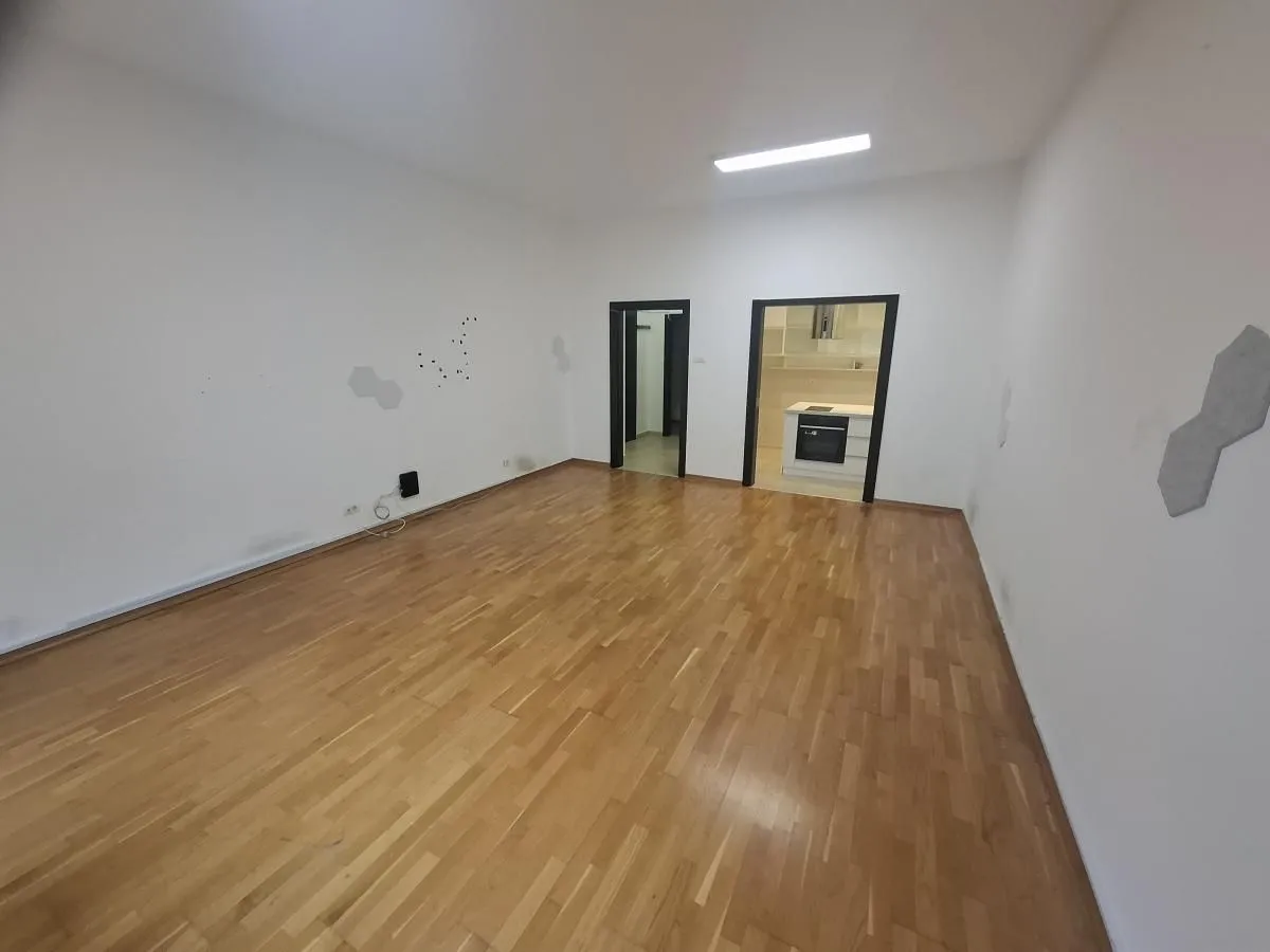 Prazna soba sa drvenim podom i pogledom na kuhinju kroz vrata.