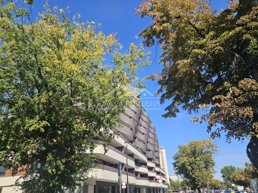 Pogled na spoljašnjost zgrade sa zelenilom i drvećem u prvom planu.
