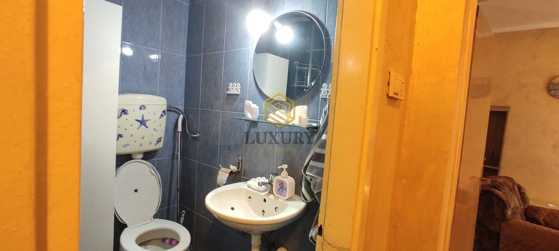 Manje kupatilo sa plavim pločicama, umivaonikom, ogledalom i WC šoljom.