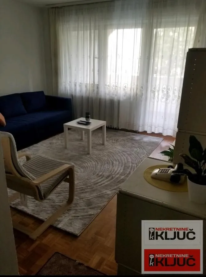 Dnevna soba sa plavom sofom, foteljom, stočićem i prozorom sa zavesama.